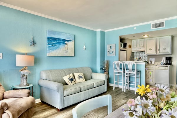 Bright open-plan beachfront condo with granite countertops, black sofa, yellow curtains, and ocean view from balcony doors