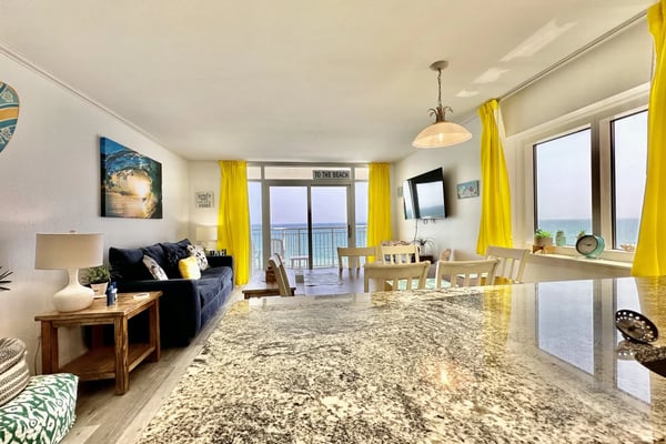 Bright open-plan beachfront condo with granite counters, navy sofa, yellow curtains, and ocean view from balcony doors.